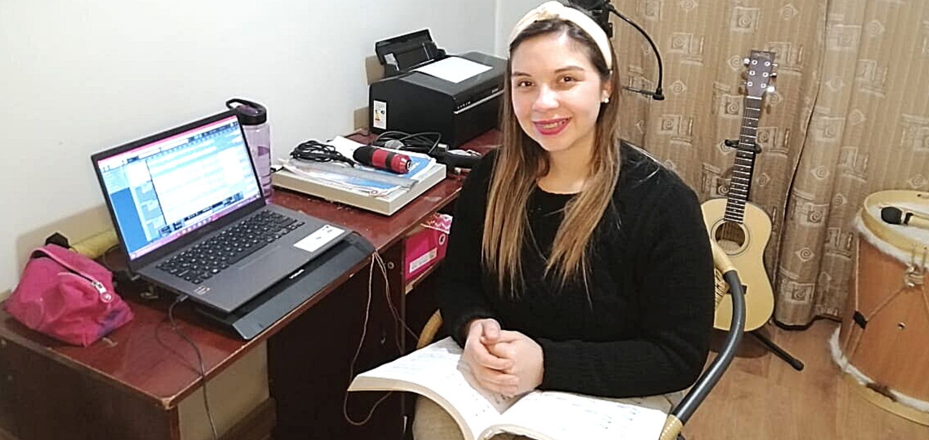 Una profesora está en su ligar de trabajo online, en donde destaca un notebook, una guitarra y sus partituras